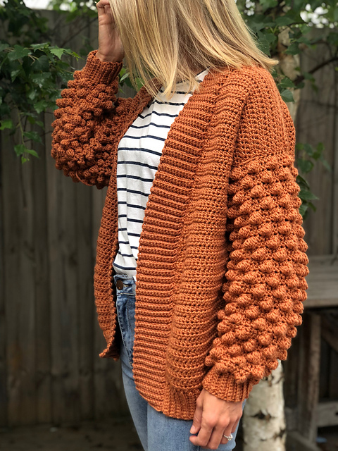 Woman wearing a rust-orange Poet crochet cardigan with oversized textured bobble sleeves, ribbed edging and a long relaxed fit.